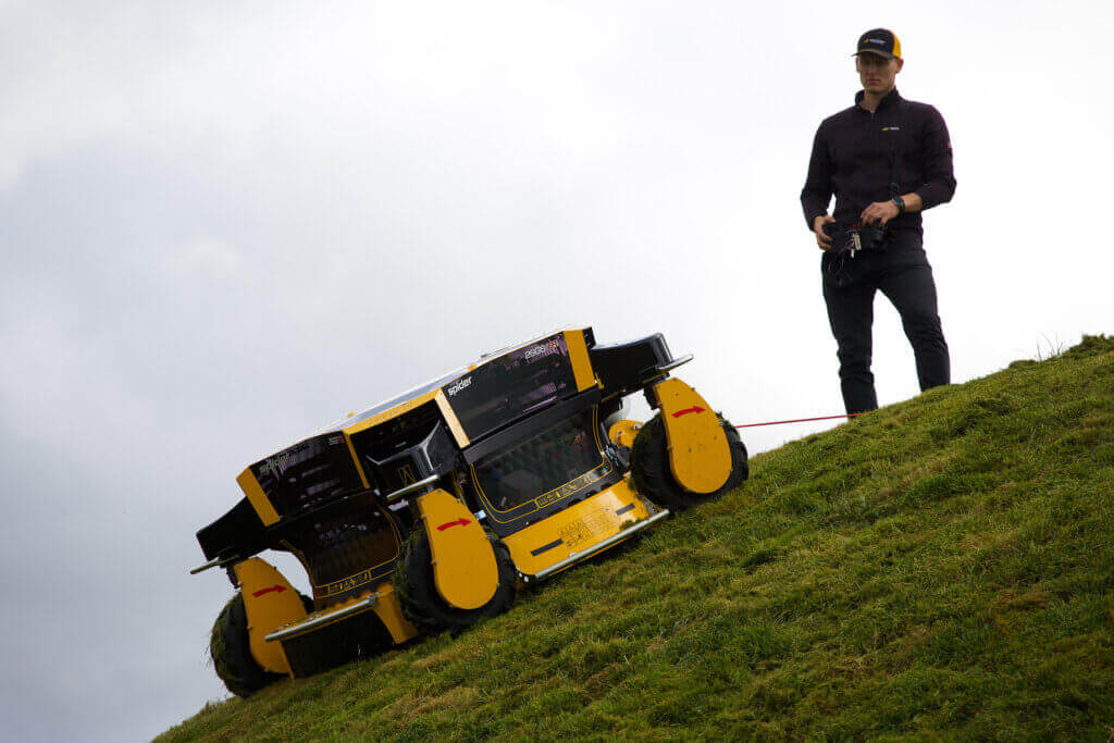 A man operating a SPIDER robotic lawn mower on sloped terrain via a remote control. 